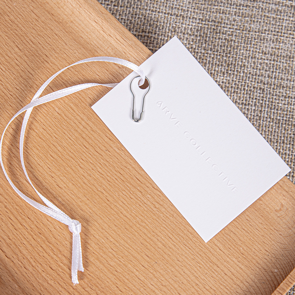 White rectangular paper hang tag with subtle impressed lettering, a white cord and a silver pin.
