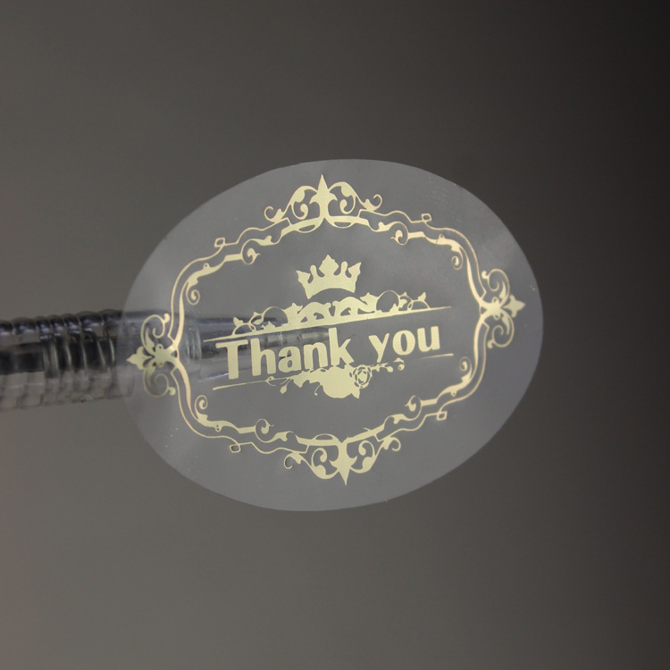 A transparent round sticker with a decorative gold-colored Thank you design.