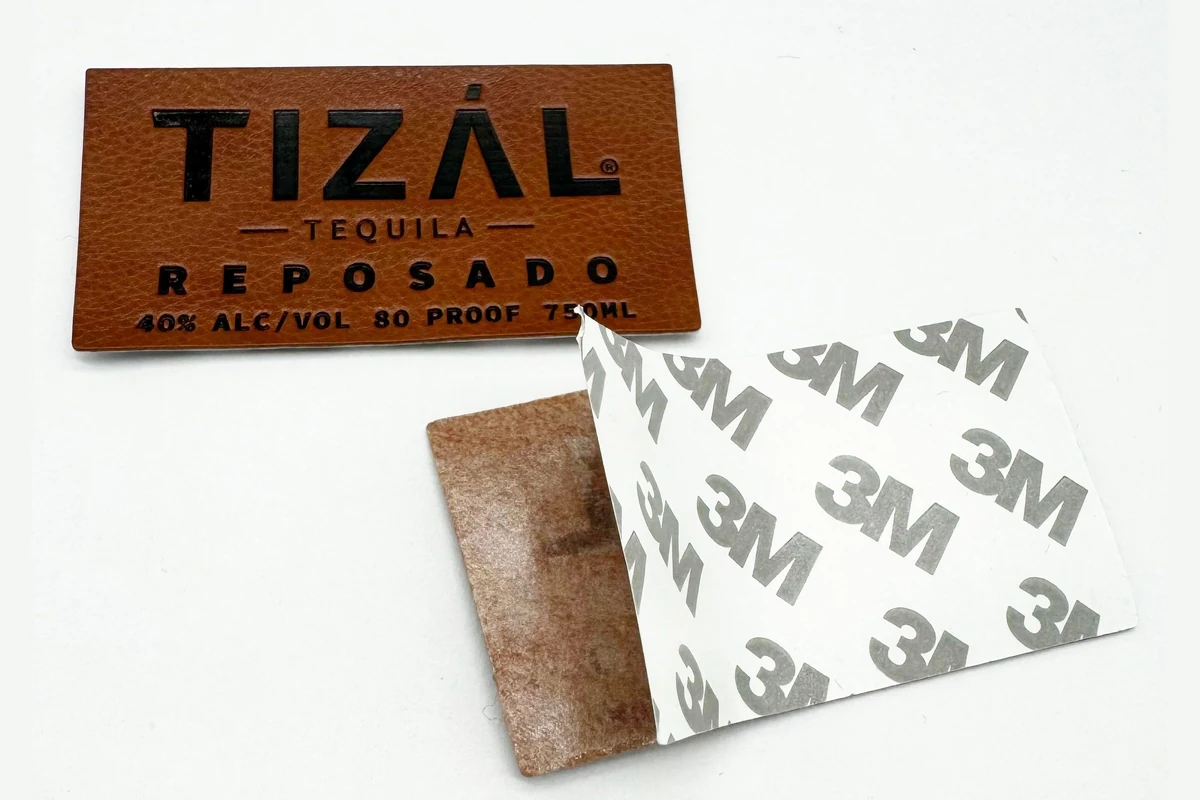 A brown printed leather label beside a sample showing adhesive backing.